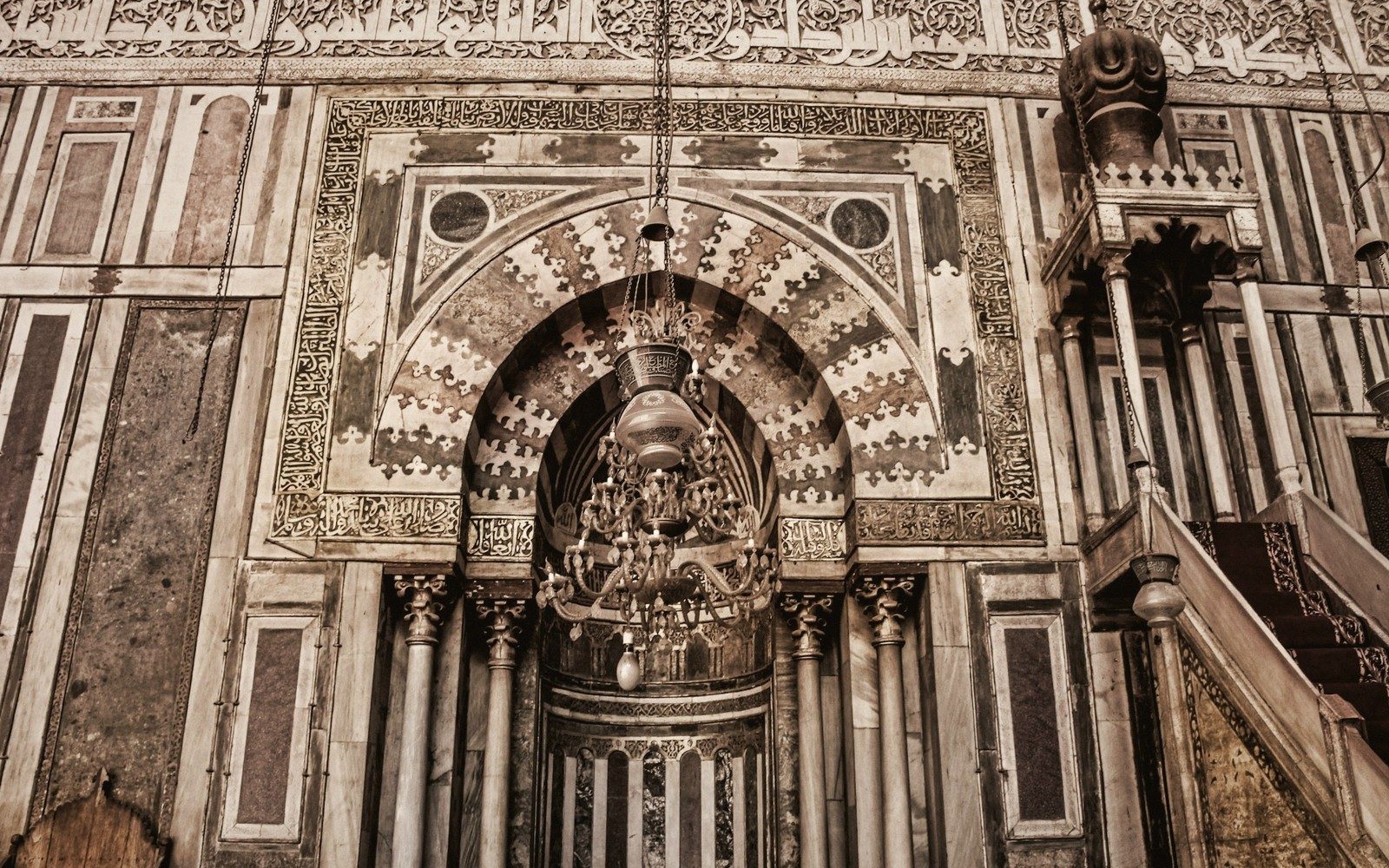 A carved marble mihrab with a hanging chandelier and adjoining minbar staircase in a mosque