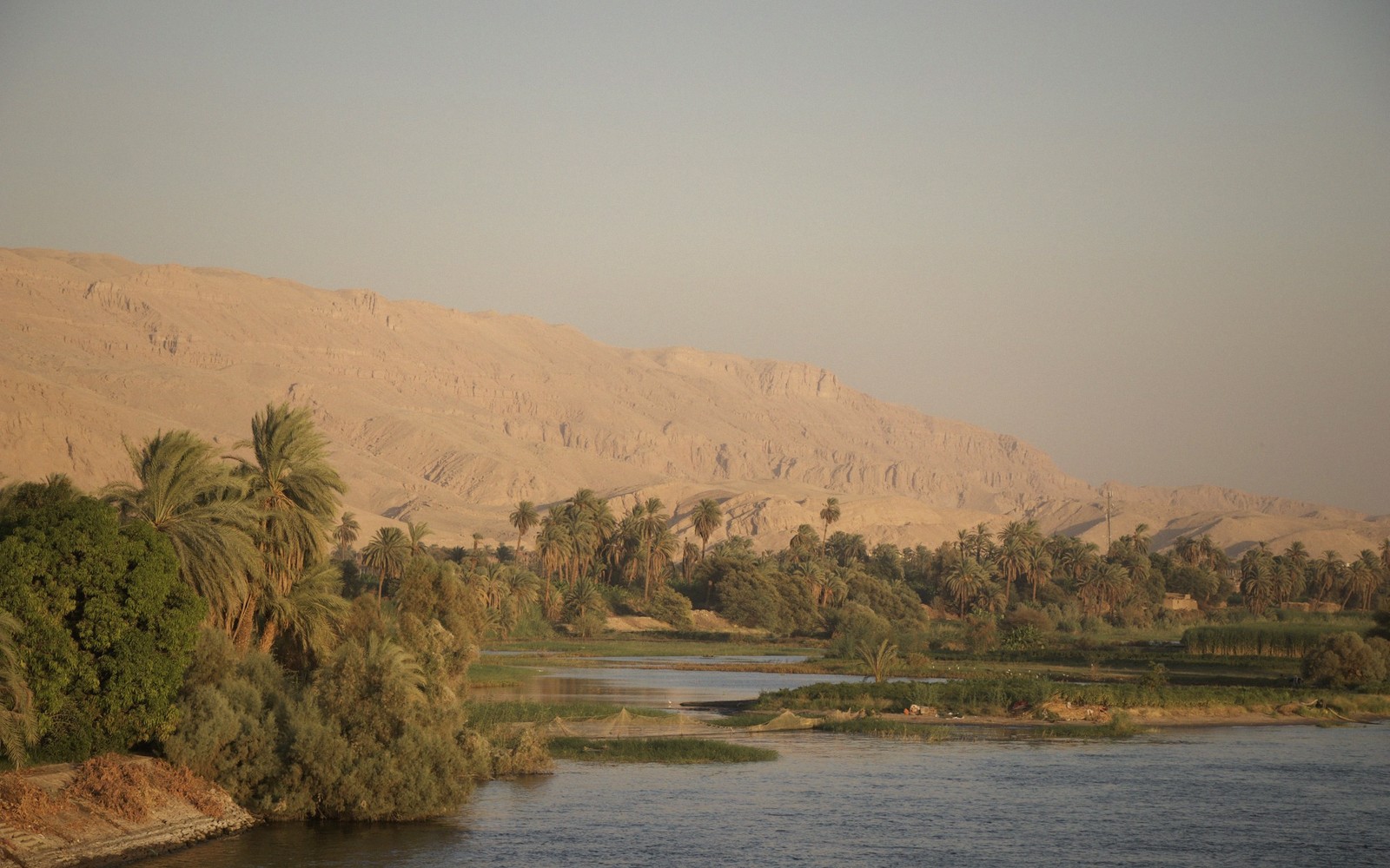 Palm trees line the Nile's banks beneath desert cliffs under a hazy sky